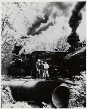 Two Shay engines with crew standing beside them at water tank along tracks on mountain trip.  