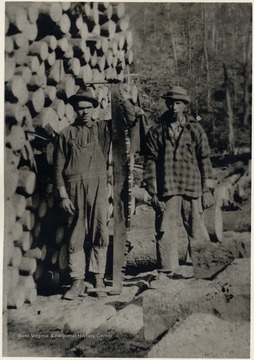 Two men pose next a pile of logs.  