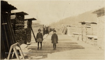 Two young girls stand among piles of lumber.