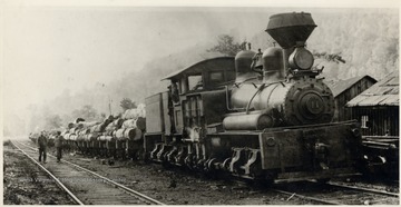 Class C Shay.  Shay train engine no. 11 pulling log carts.  Men are in the cabin.  Two people walking beside the train.