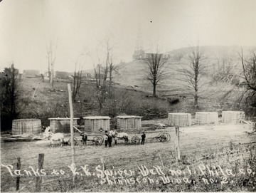 Two horse drawn carts.  Oil tanks and derrick in the background.