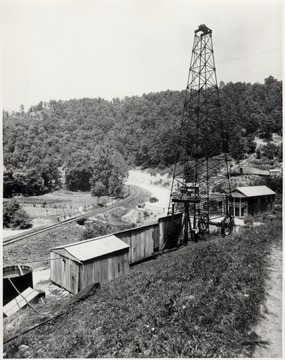 Derrick, rig, and storage tank beside a train track.