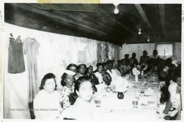 Candid group portrait of African-American Extension workers at the Filbert Achievement Day Exhibit and Luncheon. Many women are sitting at a large table with dresses hanging on the walls behind them.