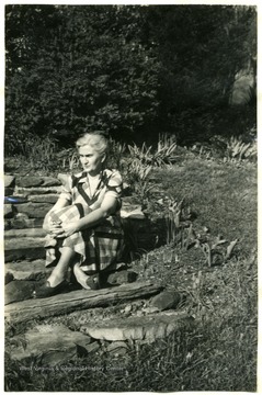 Portrait of Mrs. L.E. Griffin sitting next to her lily pool in Harrison County. Caption reads 'I built 4 foot by 9 foot by 30 inch deep pool in front of a clump of shrubbery on a bank to give it a natural setting. The bottom and sides were cemented, with a shelf built in each end for shallow water plants. From the water line the back and sides were laid up of native stones, stones also circling the front. I did most of the work herself - my husband helped dig out the last few wheelbarrows of dirt, and was home long enough to smoothe down the last of the cement. A boy helped mix the cement. As soon as it was finished I filled it with water to set till spring, using a rubber hose from a faucet in cellar - have it drained the same way. I learned from her mother that a long stick placed in a rain barrel would keep it from bursting when frozen. It also works with a lily pool. While cement was setting I planted perennials and Dutch bulbs in front of shrubbery and around the pool. It has been a mass of bloom from crocus and grape hyacinth time until now (Sept. 30) when dwarf marigolds, petunias, etc. are in their glory.'