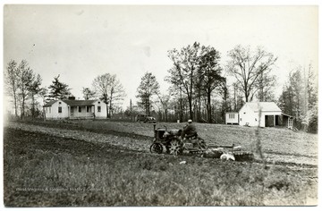 Man plowing a field with a tractor, house, car, and barn in background. 