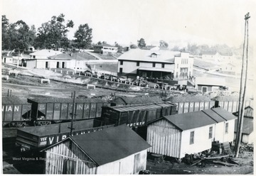 Trains next to some houses and a store with cars parked in front of it. 