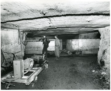 Two men stand beside rail cars.  A cart a motor sits in  front of them.