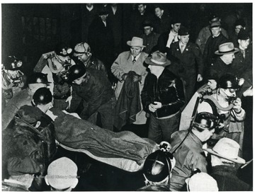 Body of a miner on stretcher in a crowd of miners in rescue gear.  Caption on back reads, 'The 112th miner was lucky, he still lives.'"