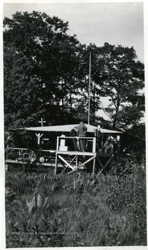 Man speaks into a microphone on a platform.  Other men standing on platform.