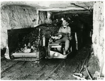 A miner is driving a battery powered shuttle car that is self unloading to the loading station. 