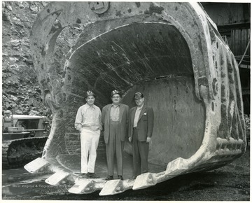 Three men stand in scoop of giant shovel. 'Bert Butler, Dia Alk.'