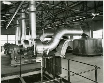 'On the top level of the heat drier building showing, from left to right, the motors, blowers and tops of the cyclones. The vertical tubes are exhaust stacks for the waste heating gases and moisture. The driers themselves are long, vertical tubes, located beneath this deck. A blast of hot gasses dries the coal and the cyclones then separate the coal from the waste gasses and moisture. Purpose of the drying operation is to reduce the moisture acquired in coal washing, 'or from the atmosphere during storage on the stock pile', and provide the processing plant with a uniform charge material.