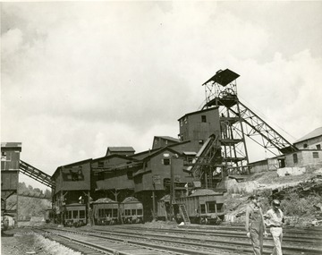 Two men walk along the tracks in front of mine buildings.