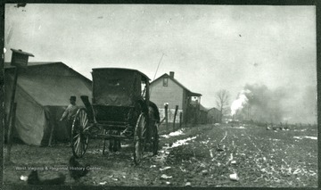 Car parked near miner's homes and a billow of smoke rising in the background.