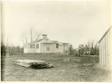 A stack of lumber sits in front of a house along with an old well with a hand pump and a car parked in the drive.