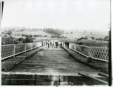 Group of men standing near the middle of the bridge. Two are fighting.  Some men on the walkways of the bridge. Edge of bridge can be seen. In the distance you can see hills and the pathway.