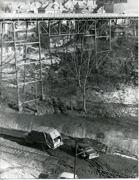 Deckers Creek and Elk Street. Morgantown and Kingwood railroad tracks. Two vehicles can be seen off of the railroad tracks. Creek at bottom near vehicles.  Neighborhood on far side of the bridge. 