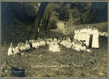 Group portrait of children attending picnic held by the Christian and Missionary Alliance Church. Children seen sitting around food. Women stand behind children on the right side of the group. 