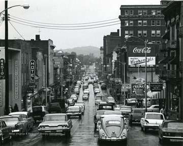 Traffic on High Street in Morgantown, W. Va. Business buildings seen along the street. Cars also parked at meters along the street. 