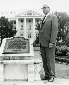 Man stands next to a memorial stone. The stone reads '1858-1922, Here stood a famous hostelry affectionately known as The Old White, once the pride of the Old Dominion, whose gracious hospitality, beautiful surroundings and healing waters gained national renown and made it the object of many a pilgrimage. Here gathered from the North and South great generals, famous statesmen and reigning belles 'who left upon the silent shore of memory images and precious thoughts that shall not die, and cannot be destroyed'. Erected by its successor The Greenbrier, 1940.'