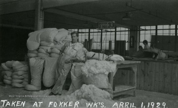 A man leans against a table at Fokker Plant while some women are seen working in the background.