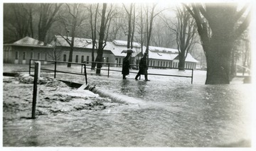 Two people walk across a bridge with flood waters flowing over it.