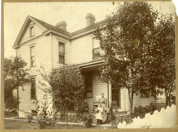 Three people are standing near the front porch of the house where Irene Burshinal Manear (wife of Clinton T. Manear) spent her young years.