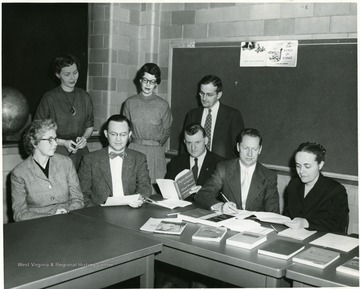 A group of individuals in a room with a display of books.  Some are seated and some are standing and engaged in a conversation.  Standing, third from left, Clifford Brown.  Seated, left to right, Grace Scott, Eston K. Feaster, rest unknown. 