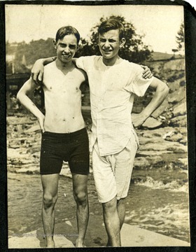 Two boys with arms on each other's shoulders near water.