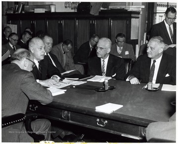 'Interstate Highway Hearings Get Under Way -- Senator Chapman Revercomb (R - WVA), seated extreme right, questions Secretary of Commerce Sinclair Weeks, at head of table, as hearings got under way this week by the Senate Roads Subcommittee on progress of the Interstate Highway System. Department of Commerce officials present for the initial session (seated clockwise) are Louis Rothschild (back to camera), under secretary for transportation; Secretary Weeks; Bradley Nash, deputy under secretary for transportation; and Bertram D. Tallamy, federal highways administrator. Reporters covering the hearing are shown in background.'