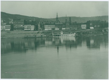 A barge and packet rest along the Ohio River shore with downtown Wheeling in the background.  