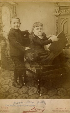 Cabinet card photograph of a small boy and girl, posed with a book. Inscribed above the image, regarding the little girl, "she became Mrs. W. S. Johnson State Treasurer [of West Virginia]".