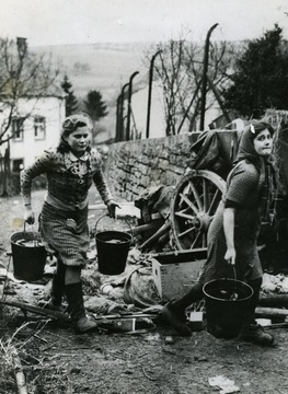 Information included on back: "Two young women of captured Rittersdorf, Germany, step over an abandoned Nazi rifle as they carry water in the Reich town, seized by troops of the Third U.S. Army February 26, 1945. The town was cleared by American soldiers driving to Bitburg, last important German road center west of the Rhine in the Third Army battle sector." 