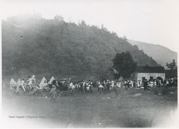 A crowd of people are scattered across a field. Parked automobiles sport American flags.