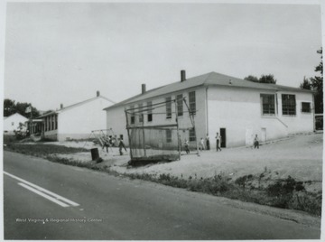 Looking at the building's exterior from across the street. Children can be seen playing by the swing-set in front of the school.