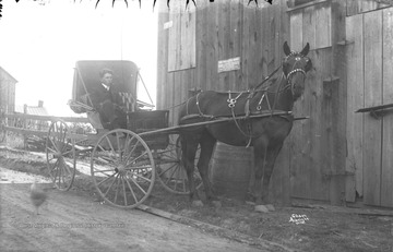 Unidentified young man with a cigar in his mouth holds the reins while parked next to a shop which carves headstones and cemetery monuments.