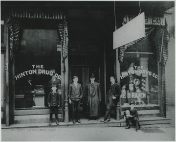 Picture of one of Hinton's early drug stores. It was located next to the First National Bank on 3rd Ave. Wade H. Gwinn, one of the store's operators, is the gentleman leaning against the post on the left. This three-story brick building housed apartments on the upper floors.