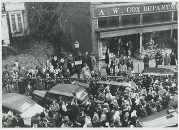 A crowd of people gather on the sidewalk and in between the parked cars. A. W. Cox Department store pictured in the background.  