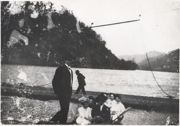 Formerly known as Pack's Ferry, A group is pictured sitting inside a small rowboat at the ferry site. Behind them, a man walks on the pier. Subjects unidentified. 