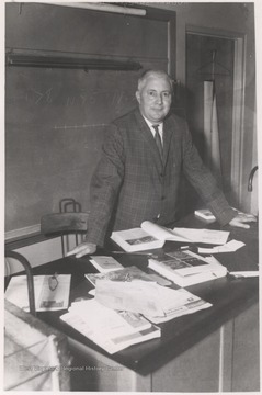 Hedrick, pictured at his book-covered desk, was a teacher at the high school. Note the fractions and number lines drawn on the chalkboard, implications that Hedrick taught mathemathics. 