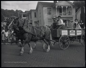 People dressed in old fashioned clothing are pulled down the street in a wagon by two horses. Signs on the cart read, "Health", "Home Improvement", and "Education." Subjects unidentified.