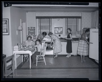 A group of women work in the kitchen while a young boy sits at a table. Subjects unidentified. 