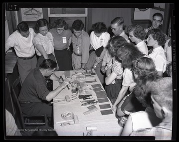 A group of young boys and girls watch as a man demonstrates how to work with leather. Subjects unidentified. 