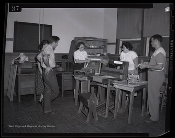 A group of young men and women watch as a women demonstrates how to weave fabric. Subjects unidentified. 