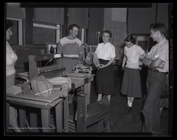 A group of young men and women listen as the instructor gives instruction on how to properly weave and design fabric patterns. 