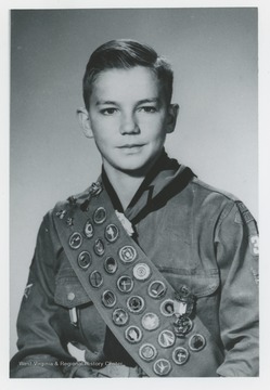 A young Koon family member in his Boy Scouts of America uniform.  Merit badges on his sash include music, camping, athletics, swimming, sports, canoeing, home repairs, first aid, cooking, firearms, and pathfinding, among others.