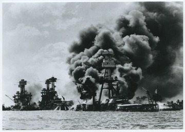 On the left, only the top deck and caged masts of the U.S.S. West Virginia can be seen. The U.S.S. West Virginia was hit with nine bombs and torpedoes total. In the center is the U.S.S. Arizona and on the right is the U.S.S. Tennessee. All ships are on fire. 