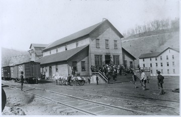 A crowd is gathered on the steps at the store's entrance. The store is located next to railroad tracks, where C. & O. railroad cars are sitting. 