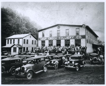 Cars are parked outside of the building where a crowd is gathered. 