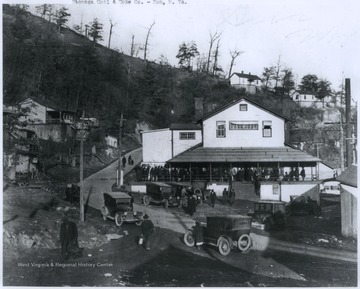 Automobiles are parked around the company building where a crowd is gathered, likely to collect paychecks.